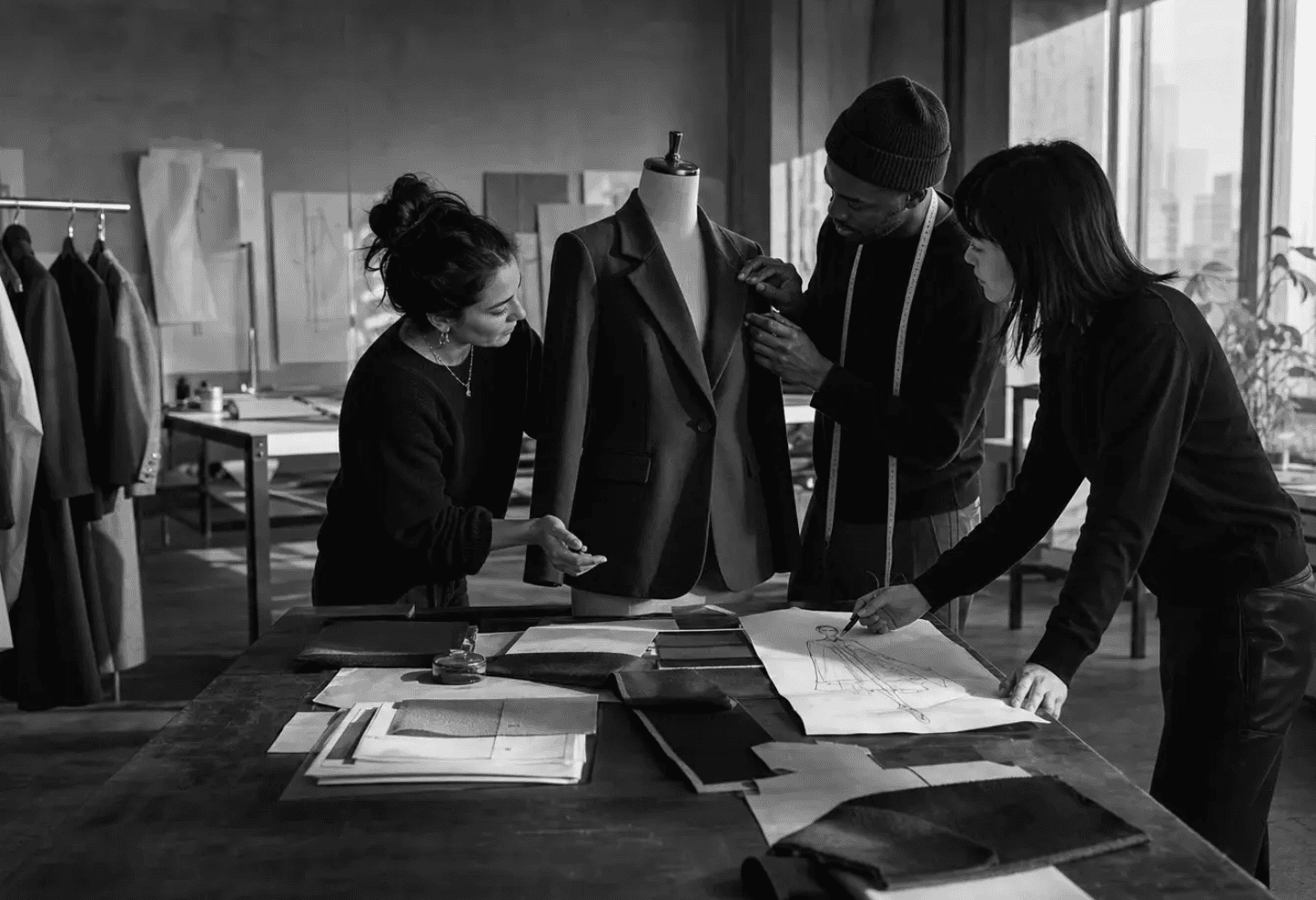 Three designers fitting a tailored jacket on a dress form beside sketches and fabric swatches